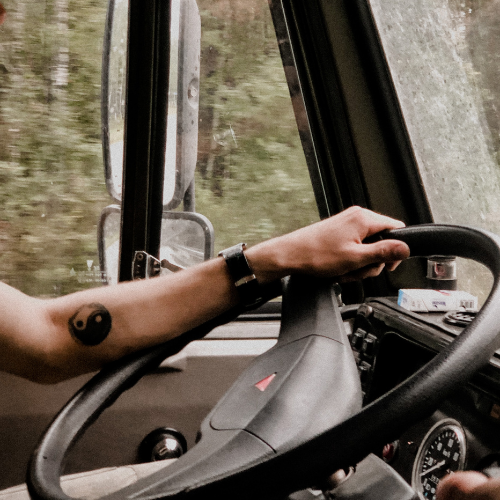 Se ve el volante de un camión. Una mano sobre el volante sugiere que un varón está conduciéndolo. Tiene un tatuaje en el antebrazo. Hay un paisaje verde detrás
