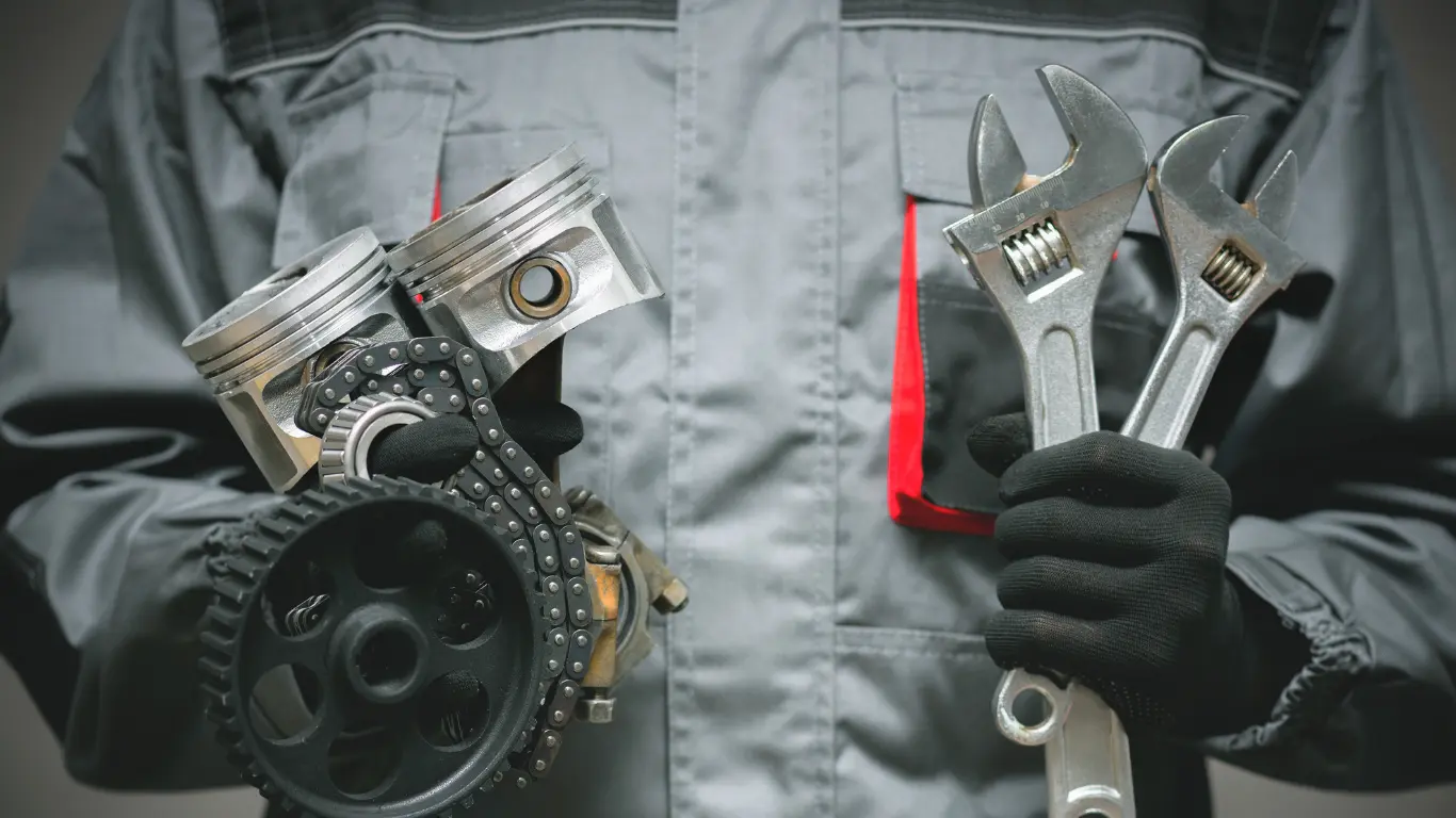 Pecho de un mecánico, de guantes negros. En la mano izquierda tiene correas y pistones y en la mano derecha llaves inglesas.